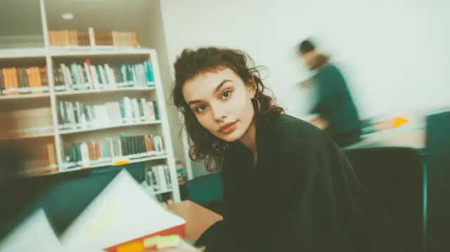 Młoda kobieta w okularach siedzi przy biurku w bibliotece, otoczona książkami.