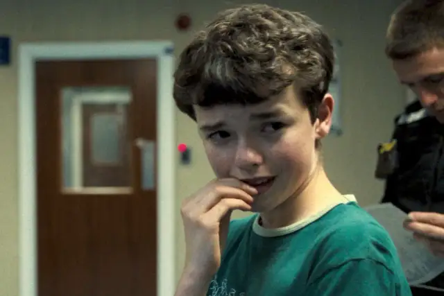 Chłopiec zamyślony, jakby pochłaniał wszechświat w swoich myślach, z ręką przy ustach. W tle drzwi i postać w mundurze.