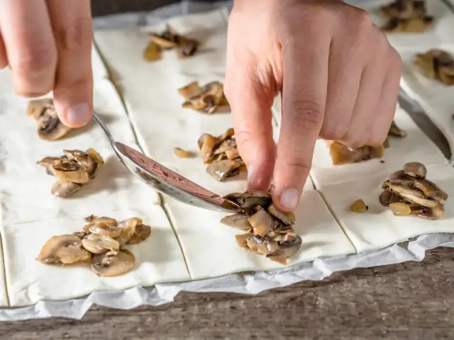 Ręce układają pieczarki na cieście francuskim, przygotowując pyszne przekąski.