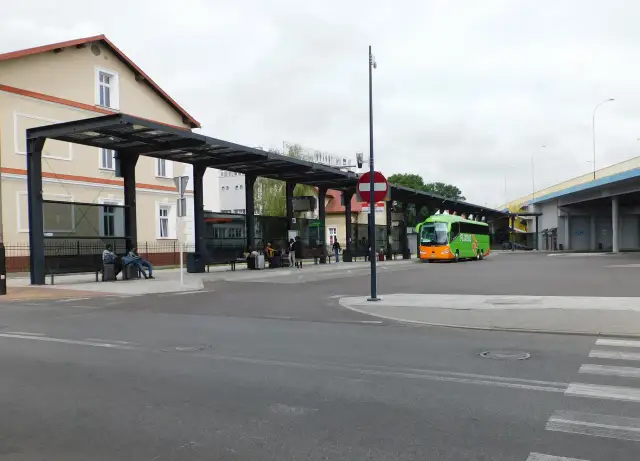 Zielony autobus FlixBus na przystanku autobusowym z wiatą i ławkami, gdzie siedzą pasażerowie.