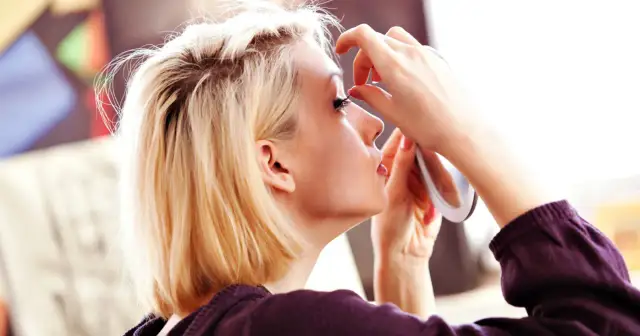 Kobieta z blond włosami przygląda się w lusterku, dotykając palcem rzęs.