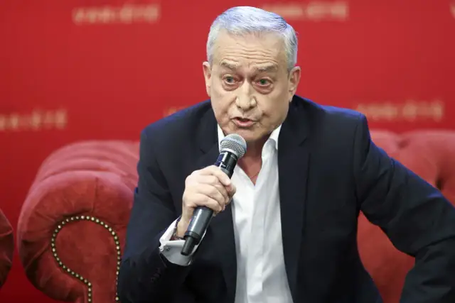 Janusz Szydłowski przemawia do mikrofonu, siedząc na czerwonej, pikowanej kanapie.