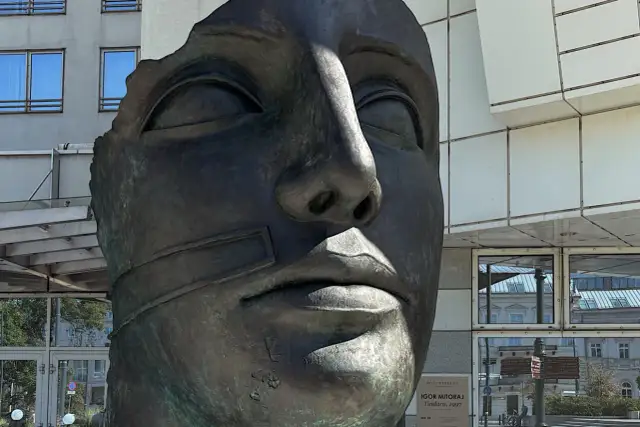 Brązowa, fragmentaryczna rzeźba twarzy Igora Mitoraja na tle nowoczesnego budynku.