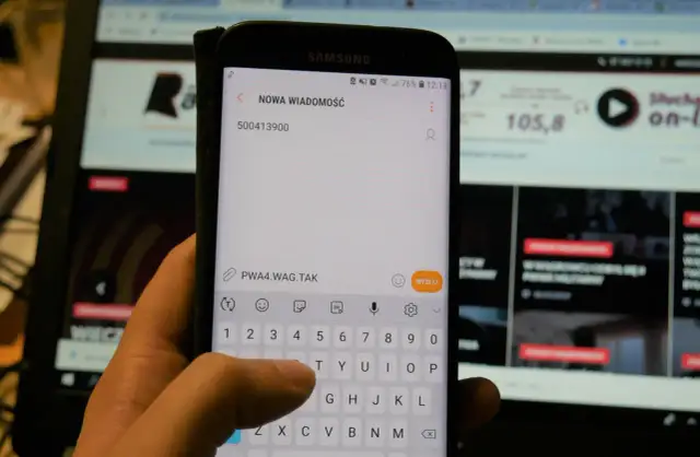 Dłoń pisze SMS na telefonie z numerem 500413900. W tle ekran komputera.