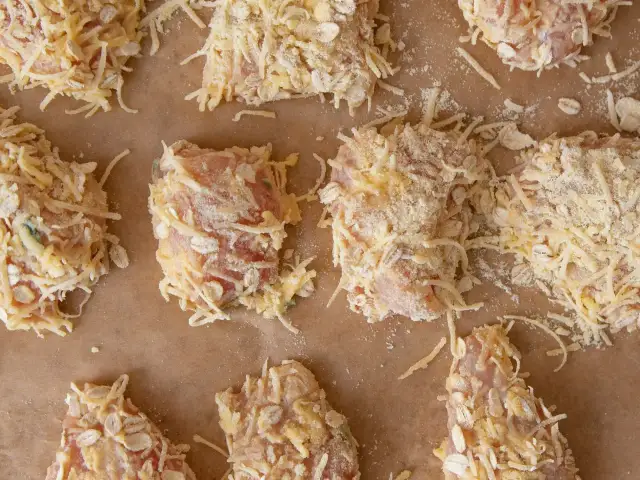 Kawałki fileta z kurczaka obtoczone w tartej bułce, serze i płatkach owsianych, ułożone na papierze do pieczenia.