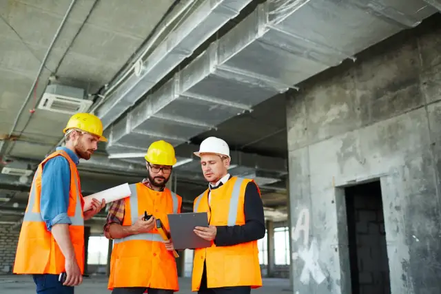 Trzech budowlańców w kaskach i kamizelkach omawia dokumenty na placu budowy.