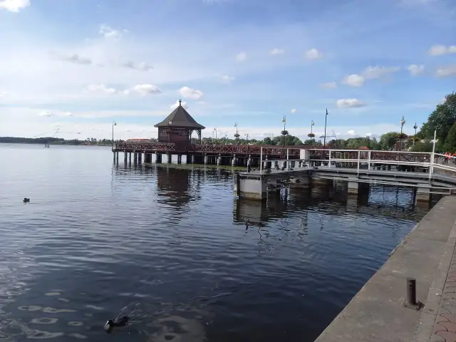 Drewniana altana na molo w Ostródzie, otoczona wodą i zielenią. Kaczki pływają spokojnie.