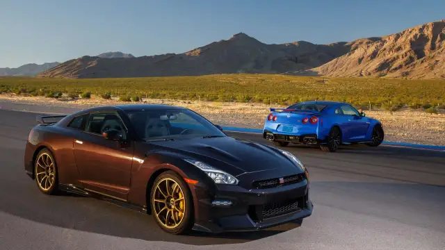 Dwa Nissan GTR R35, jeden brązowy, drugi niebieski, stoją na torze wyścigowym na tle pustynnego krajobrazu.