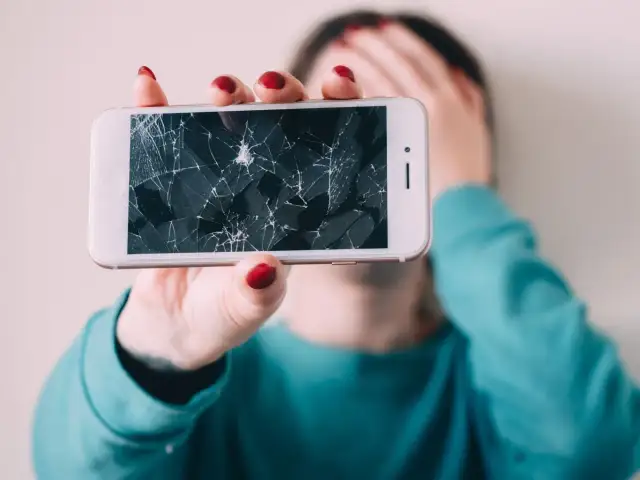 Złamany telefon z pękniętym ekranem, który osoba trzyma przed twarzą, zakrywając ją ręką.