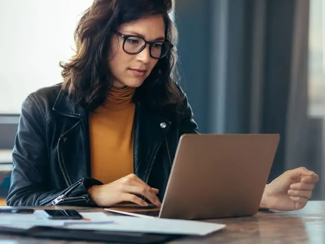 Kobieta w okularach i skórzanej kurtce pracuje na laptopie, szukając informacji, jak sprawdzić lata pracy w ZUS PUE.
