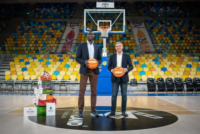 Dos hombres posan con balones de baloncesto en una cancha vacía. Detrás de ellos, una pila de cajas de frutas y asientos de colores. Un guiño al club baloncesto Gran Canaria.