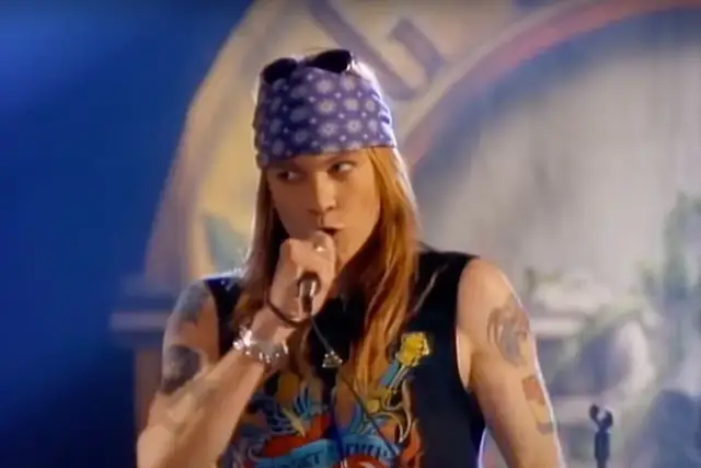 Axl Rose w fioletowej bandanie i czarnej koszulce śpiewa do mikrofonu, jakby dedykował "sweet child o' mine" publiczności.