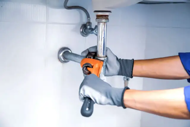 Hydraulik w rękawiczkach dokręca kluczem instalację pod zlewem.