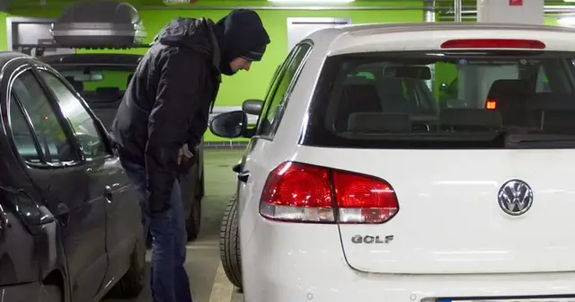 Mężczyzna w kapturze pochyla się nad białym Volkswagenem Golfem, jednym z najczęściej kradzionych aut.