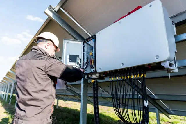 Technik w białym kasku sprawdza instalację fotowoltaiczną. Falownik pokazuje błąd, co wymaga jego uwagi i naprawy.
