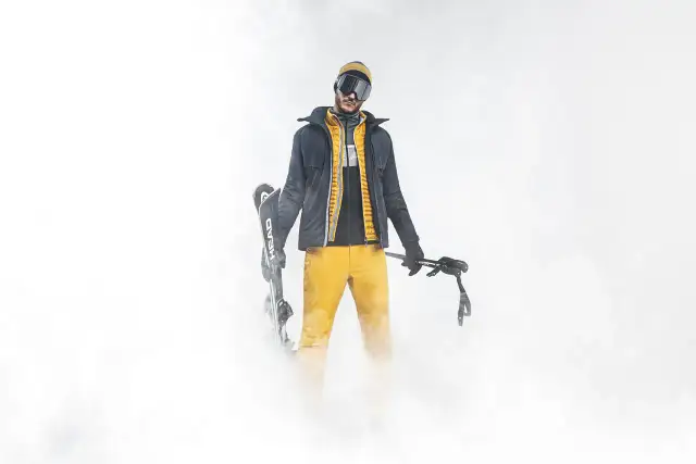 Mężczyzna w żółtych spodniach narciarskich i ciemnej kurtce, trzymający narty i kije. Idealny przykład, jak dobrać spodnie narciarskie.