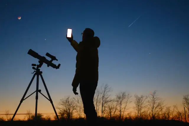Sylwetka osoby z telefonem i teleskopem na tle nieba z zaćmieniem Księżyca i smugą po samolocie.