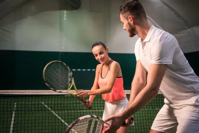 Para z rakietami tenisowymi na korcie, mężczyzna uczy kobietę gry.