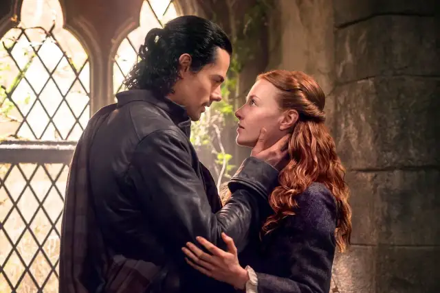Mężczyzna z czarnymi włosami czule dotyka policzka kobiety o rudych włosach. Scena z serialu "Outlander: Krew z krwi".