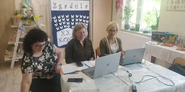 Trzy kobiety pracują przy stołach w sali, która wygląda jak szkoła specjalna. Na ścianie wisi tablica z motylami i napisem "Zgadula".