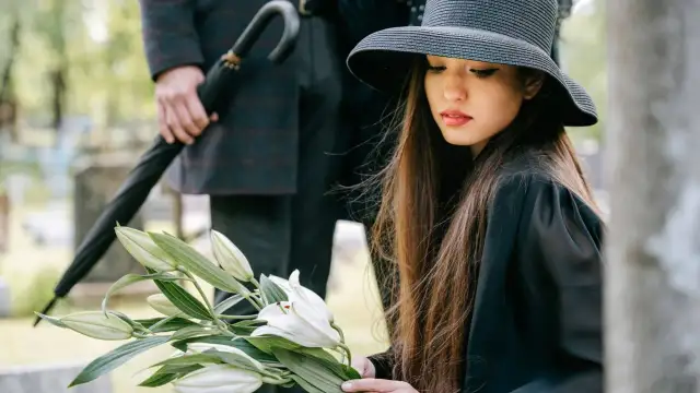 Kobieta w czarnym kapeluszu trzyma bukiet białych lilii na cmentarzu.