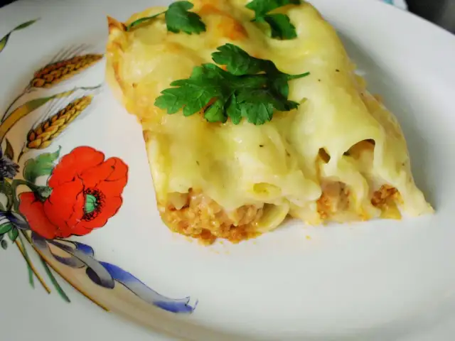 Cannelloni z mięsem i beszamelem przepis. Pyszne danie zapieczone pod beszamelem, udekorowane natką pietruszki, na talerzu z makami.
