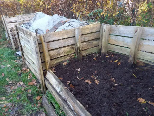 Drewniany kompostownik w ziemi, częściowo przykryty plandeką. Widać jesienne liście i trawę.