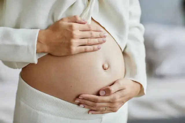 Zbliżenie na ciążowy brzuch kobiety, która delikatnie go obejmuje dłońmi.