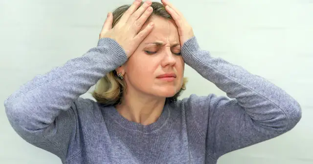 Kobieta z rękami na skroniach, z zamkniętymi oczami i grymasem bólu na twarzy.
