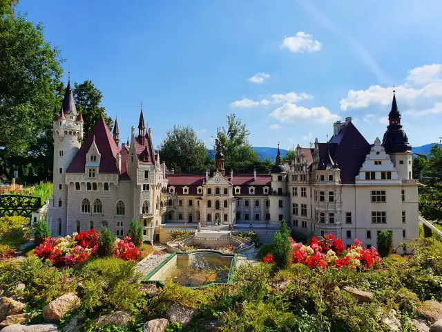 Miniatura zamku z ogrodem i fontanną, otoczona kwitnącymi kwiatami.