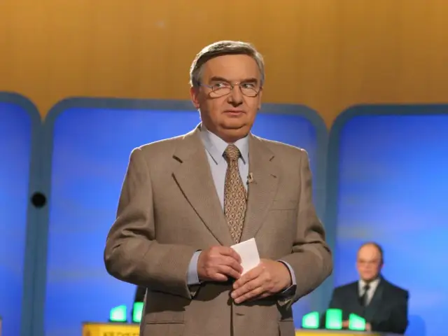 Tadeusz Sznuk, który prowadzi teleturniej Jeden z dziesięciu, w beżowej marynarce i z kartką w dłoni.