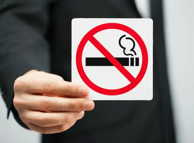 Dłoń w garniturze trzyma tabliczkę z zakazem palenia.