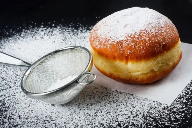Puszysty pączek posypany cukrem pudrem, obok sitko z cukrem. Idealne na tłusty czwartek, pyszne pączki!