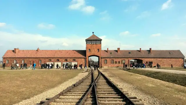 Brama obozowa Auschwitz-Birkenau z torami kolejowymi i grupą zwiedzających.