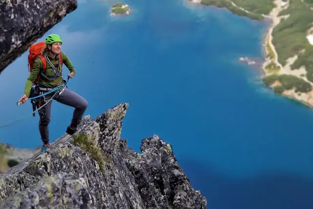 Kobieta w kasku i sprzęcie wspinaczkowym na skale, z widokiem na turkusowe jezioro. To idealne miejsce na kurs taternicki.