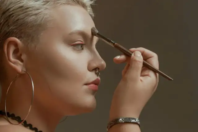 Kobieta z krótkimi blond włosami i kolczykiem w nosie nakłada bronzer na czoło, ucząc się, jak wyszczuplić twarz bronzerem.