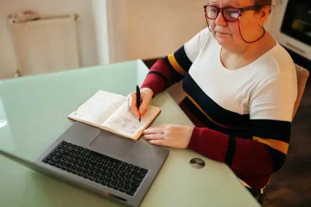Kobieta w okularach pisze w notatniku obok laptopa.