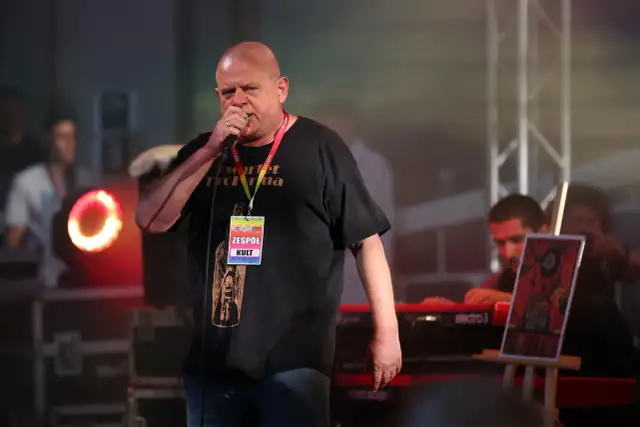 Kazik Staszewski śpiewa na koncercie, ubrany w czarną koszulkę z napisem "Kwartet Pro Forma" i identyfikatorem "Zespół KULT".