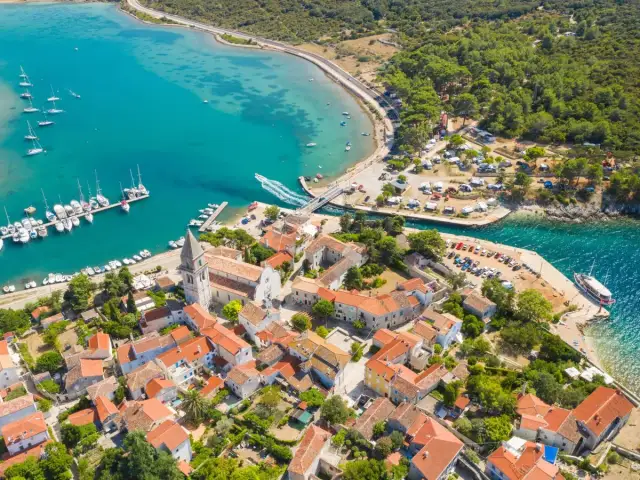 Widok z lotu ptaka na chorwacką wioskę z czerwonymi dachami, port z łodziami i turkusowe morze.