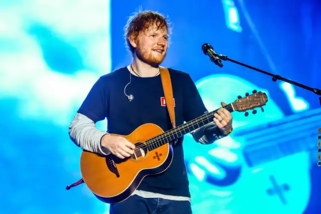 Ed Sheeran gra na gitarze akustycznej na tle niebieskiej sceny.
