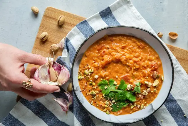 Aksamitna zupa z soczewicy z mlekiem kokosowym, posypana pistacjami i kolendrą. Obok czosnek i orzechy.