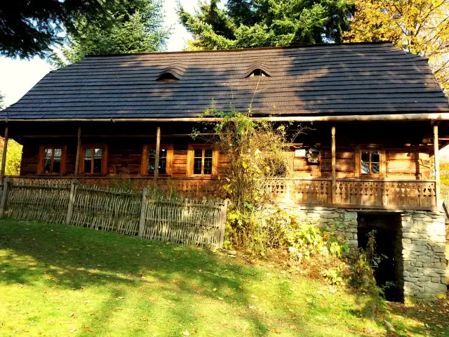 Drewniany dom z kamienną piwniczką i ozdobną werandą, otoczony zielenią i jesiennymi liśćmi.