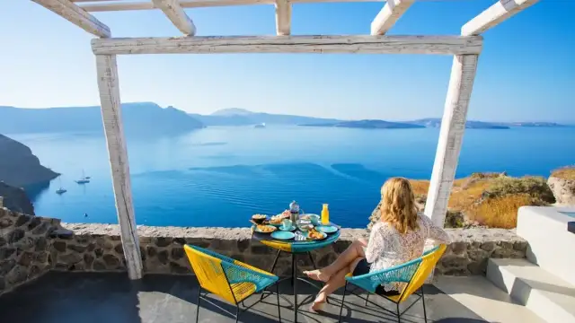 Mujer desayunando en balcón con vista al mar azul y montañas.