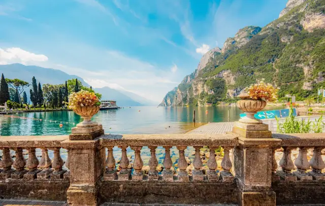 Kamienna balustrada z wazonami kwiatów przed widokiem na największe jezioro we Włoszech, otoczone górami i błękitnym niebem.