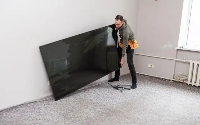 Mężczyzna w pasie narzędziowym trzyma duży, czarny telewizor przy ścianie.