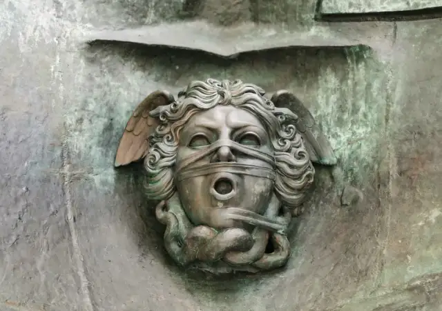 Brązowa rzeźba głowy z opaską na oczach i ustach, ze skrzydłami i wężami.