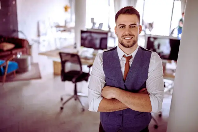 Uśmiechnięty mężczyzna w kamizelce i krawacie stoi ze skrzyżowanymi rękami w nowoczesnym biurze.