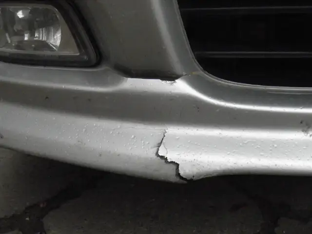 Pęknięty srebrny zderzak samochodu z widocznymi odpryskami lakieru i kroplami wody.