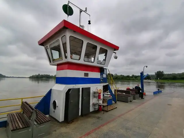 Prom z pomostem, kabiną kapitańską i ławkami na tle rzeki i zachmurzonego nieba.
