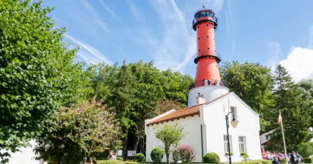 Czerwona latarnia morska w Jastrzębiej Górze na tle zieleni i błękitnego nieba.